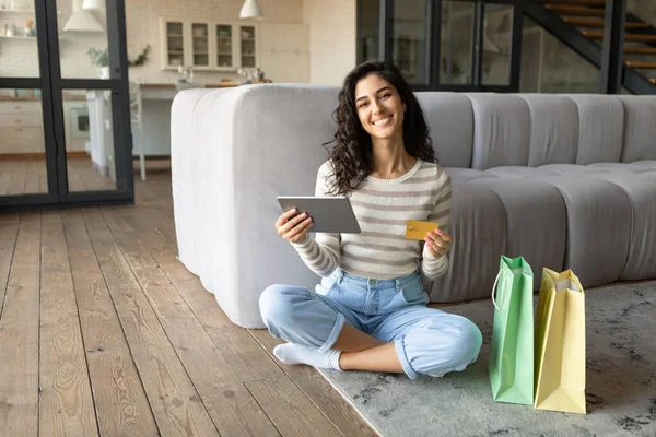 Feliz joven morena con tableta digital y bolsas de regalo con tarjeta de crédito mientras compra en línea desde casa — Foto de Stock