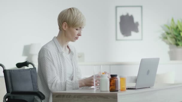 Mujer madura con discapacidad que toma pastillas y agua potable, sentado en silla de ruedas y la lectura cita en el ordenador portátil — Vídeos de Stock