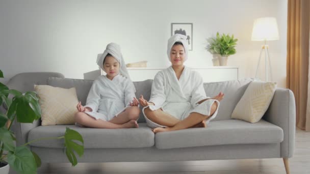 Азиатская мать и дочь в халатах и полотенцах на головах медитируют, сидят в позе лотоса на диване, увеличивают масштаб — стоковое видео