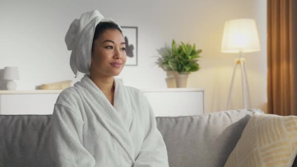 Mulher asiática bonita em roupão de banho com toalha na cabeça sorrindo para a câmera, descansando em casa após os procedimentos de banho, zoom em — Vídeo de Stock