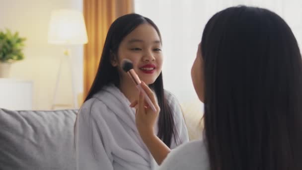 Zoom no retrato de pequena menina asiática com maquiagem profissional sorrindo para a mãe, amando a mãe aplicando rouge à filha — Vídeo de Stock