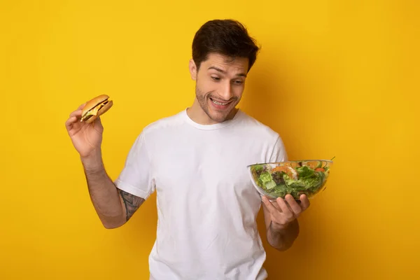 Retrato de un joven divertido sosteniendo hamburguesa y ensalada —  Fotos de Stock