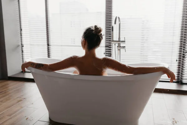 Vue arrière de la jeune femme prenant une baignoire mousseuse, regardant par la fenêtre dans la salle de bain moderne à la maison, espace vide — Photo