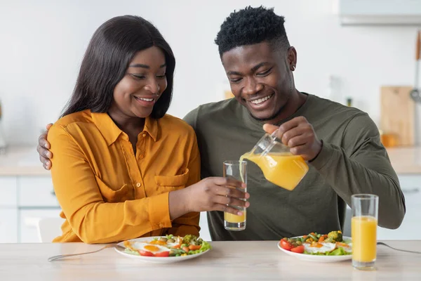 Feliz joven negro cónyuges desayunando juntos, marido vertiendo jugo de la jarra — Foto de Stock