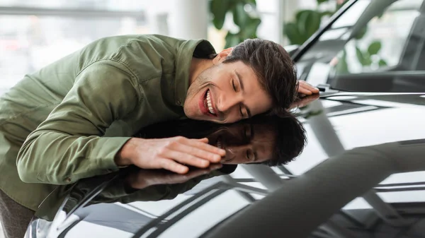 Glücklicher junger Mann umarmt Motorhaube seines neuen Autos, träumt vom Autokauf im Autohaus, Panorama — Stockfoto