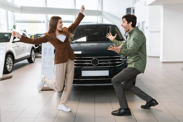 Jovem casal caucasiano animado comemorando a compra bem sucedida do carro, dançando e se divertindo na concessionária de automóveis — Fotografia de Stock