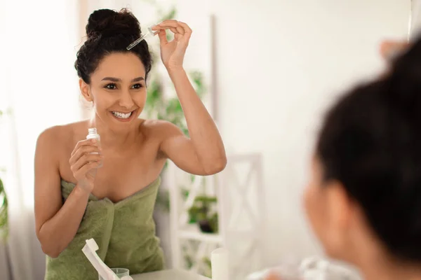 Sorrindo jovem morena em toalha aplicando soro ou óleo natural no cabelo seco danificado perto do espelho em casa — Fotografia de Stock