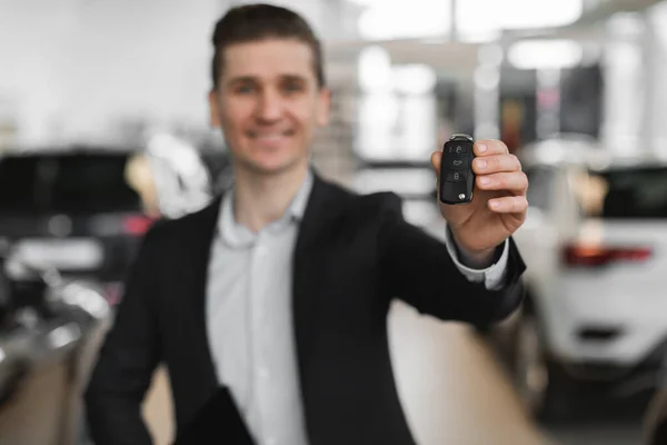 Confident young salesman showing car key at camera in modern dealership, selective focus — Stock Photo, Image