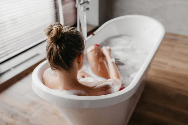 Vue arrière de la jeune femme prenant une baignoire mousseuse tout en étant couché dans la baignoire dans la salle de bain moderne intérieur à la maison — Photo