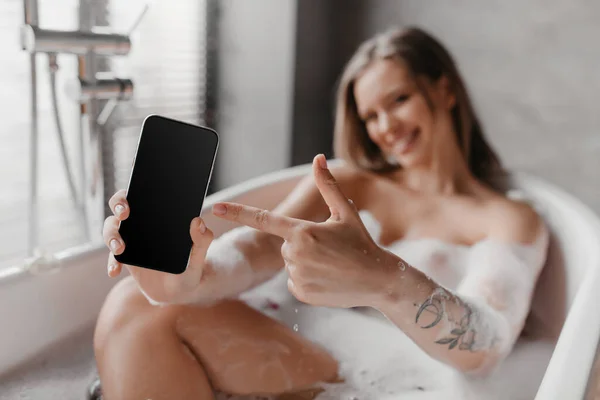 Дівчина тримає смартфон і вказує на порожній екран, показуючи рекламу, розслабляючись у пінистій ванні, вибірковий фокус, макет — стокове фото