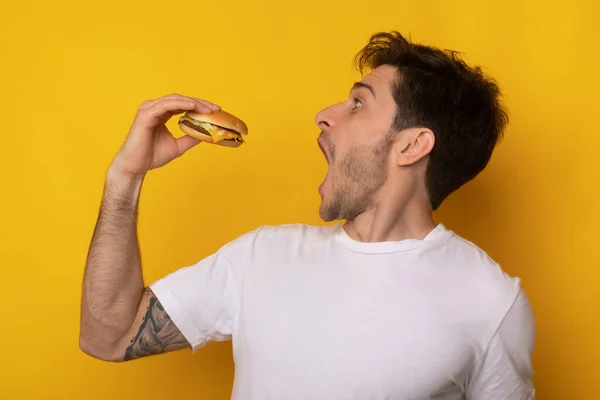 Cara engraçado segurando hambúrguer mordendo sanduíche no estúdio — Fotografia de Stock