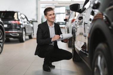 Mutlu genç araba satıcısı yeni otomobilin yanında oturuyor, kontrol ediyor, otomobil galerisinde notlar alıyor.