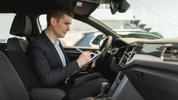 Jeune vendeur d'automobiles assis à l'intérieur de la voiture neuve, faire un check-up, écrire des données chez un concessionnaire moderne, panorama — Photo