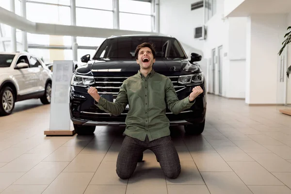 Izgatott fiatalember kiabál, rázza az öklét, ünnepli az új autó vásárlását, térden állva áll a kereskedésben. — Stock Fotó