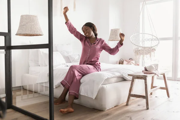 Välsovande svart dam stretching armar bär Pajamas sitter i sovrummet — Stockfoto