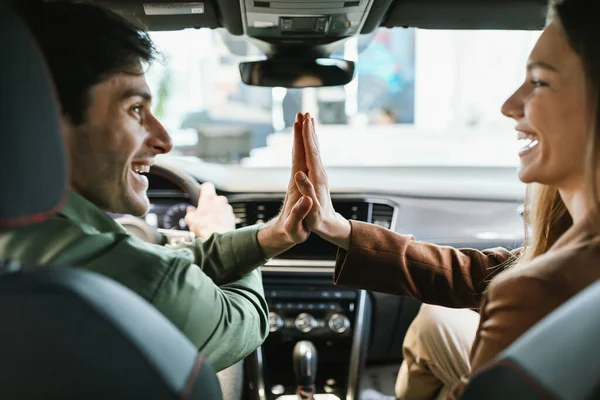 Spännande unga makar firar köp av ny bil, sitter inne i modern bil, hög femma till varandra på återförsäljaravtal — Stockfoto