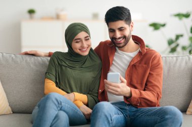 Orta Doğulu çift evdeki koltukta oturan cep telefonunu kullanıyor.