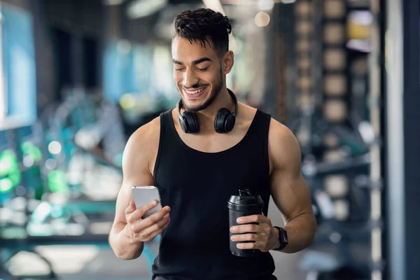 Atleta maschio arabo sorridente che si rilassa con Smartphone dopo l'allenamento in palestra — Foto Stock