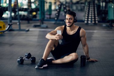 Fitness Uygulaması. Gülümseyen Orta Doğulu Sporcu Spor Salonunda Akıllı Telefon Kullanıyor