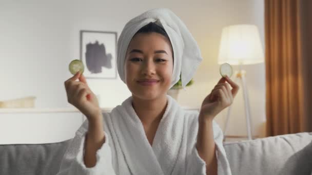 Zoom en retrato de mujer asiática bonita en albornoz con toalla en la cabeza poniendo rodajas de pepino fresco a los ojos, cámara lenta — Vídeos de Stock