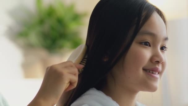 Догляд за волоссям для дітей. Мила маленька азіатська дівчинка посміхається, її любляча мати розчісує волосся в домашньому інтер'єрі, повільний рух — стокове відео