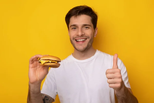 Divertido chico sosteniendo hamburguesa mostrando pulgar hacia arriba — Foto de Stock