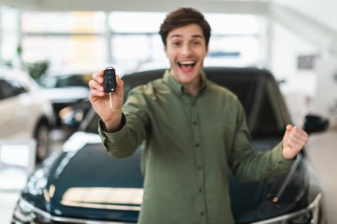 Heyecanlı genç beyaz adam yeni araba anahtarını kameraya gösteriyor, EVET işareti yapıyor, galeride araba alımını kutluyor.
