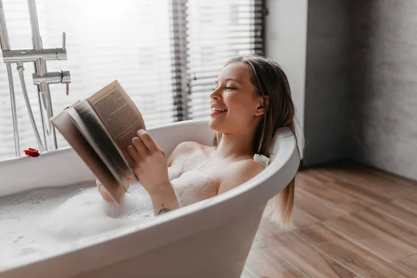 Mujer feliz tumbada en un baño espumoso, leyendo libro en un ambiente relajante, disfrutando de una historia interesante en la bañera en casa —  Fotos de Stock