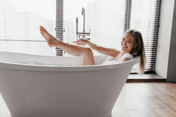 욕조에서 휴식을 취하고 발을 면도 하며 안전 한 면도칼을 사용하여 카메라를 보고 미소짓는 행복 한 여인 — 스톡 사진