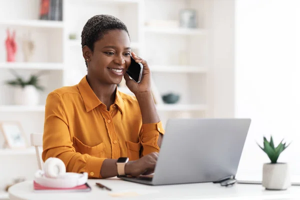 Donna nera seduta a tavola con computer portatile, avendo conversazione telefonica — Foto Stock
