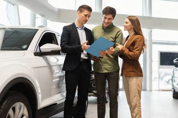 Confident car salesman showing purchase details to young Caucasian couple at dealership center — Stock Photo, Image