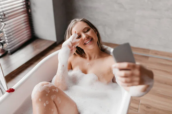 Vidám nő fekszik habos fürdő és szelfi okostelefon, pihentető és élvezi a szépség reggeli rutin — Stock Fotó