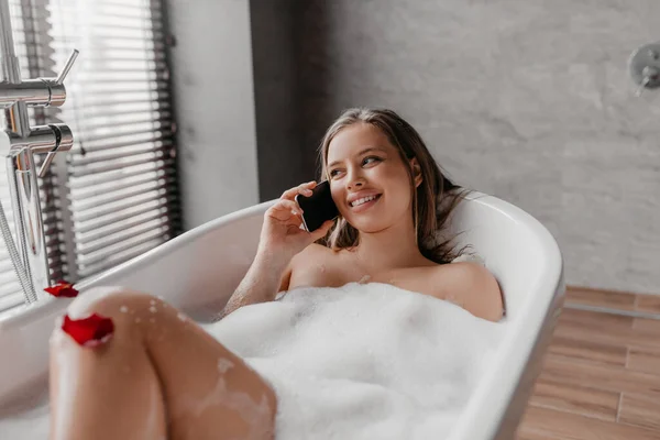 Boldog szép hölgy fekszik habos fürdő és beszél az okostelefon, pihentető fürdőkád hétvégén, szabad hely — Stock Fotó