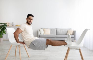 Atletik genç Arap adam evinde iki sandalyeyle güç egzersizleri yapıyor.