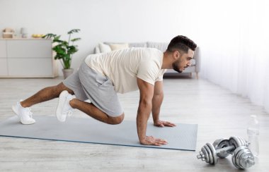 Spor kıyafetli yakışıklı bir Arap, yoga minderiyle egzersiz yapıyor, ev içi spor yapıyor, kaslarını evde çalıştırıyor.