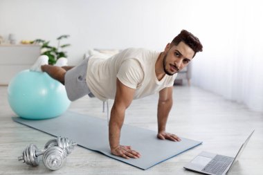 Genç Arap adam dizüstü bilgisayarın yanında duruyor, şınav için fitness topunu kullanıyor, online spor dersleri veriyor.