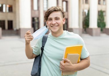 Mutlu öğrenci maskesini çıkarıyor, sırt çantası ve kitaplarla dışarıda poz veriyor, üniversite binasının yanında duruyor.