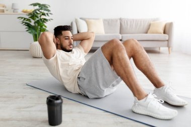 Tam boy spor yapan genç bir Arap, vücut kaslarını güçlendiriyor, evde karın kasları egzersizi yapıyor.