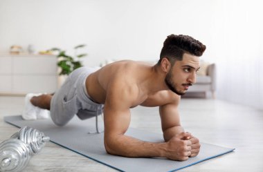 Gömleksiz genç Arap adam yoga minderinin üzerinde dirsek tahtasında duruyor, iç mekanda kas egzersizi yapıyor.
