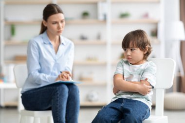 Küçük çocuk kollarını kavuşturmuş, danışmanlık sırasında kadın psikologla iletişimi ihmal etmiş.