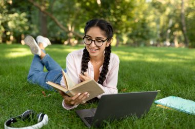 Gözlüklü mutlu Hintli kız dışarıda kitap okuyor, sınavlara hazırlanıyor ve parkta ders çalışmaktan keyif alıyor.
