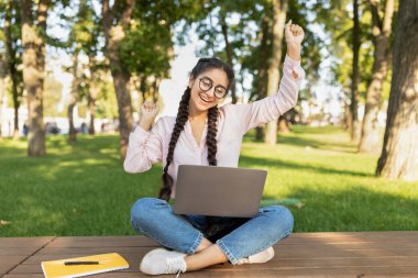 Laptop bilgisayarlı aşırı neşeli Hintli kız öğrenci açık havada EVET işareti yapıyor, üniversite kampüsünde oturuyor.