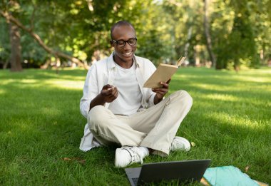 Sınav çalışma konsepti. Mutlu Afro-Amerikan öğrenci kitap okuyor ve dışarıda dizüstü bilgisayar kullanıyor.