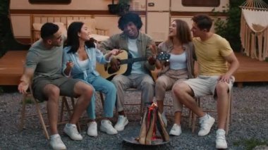 Şenlik ateşinin etrafında oturmuş gitar çalıp şarkı söyleyen çok ırklı arkadaşlar grubu.
