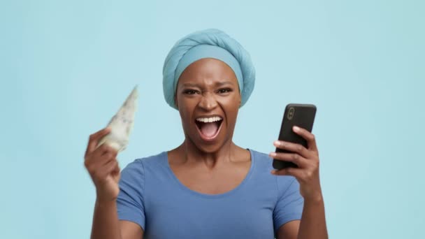Vzrušený Afroameričanka drží mobilní telefon a peníze, modré pozadí — Stock video