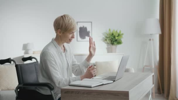 Віртуальна ділова зустріч. Щаслива жінка середнього віку користувач інвалідного візка махає рукою до ноутбука, вітаючи своїх партнерів онлайн — стокове відео