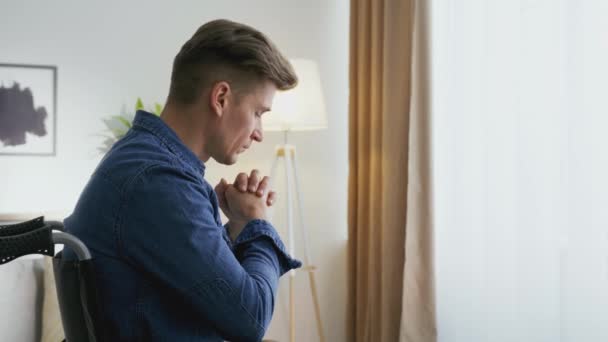 Збільшити зображення безнадійного середнього чоловіка з інвалідністю молячись до Бога, сидячи в кріслі на колесах біля вікна — стокове відео
