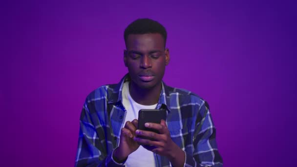 Σοβαρός μαύρος τύπος που στέλνει μηνύματα χρησιμοποιώντας το κινητό τηλέφωνο πάνω από μωβ φόντο — Αρχείο Βίντεο