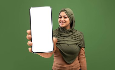 Beyaz ekranlı akıllı telefonun görüntüsü tesettürlü mutlu Müslüman kadının ellerinde, yeşil arkaplan