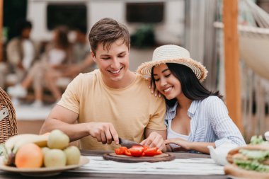 Genç adam ve Asyalı kız arkadaşı kamp gezisinde birlikte yemek pişiriyor, karavanın yanında sebze kesiyor, dışarıda vakit geçiriyorlar.